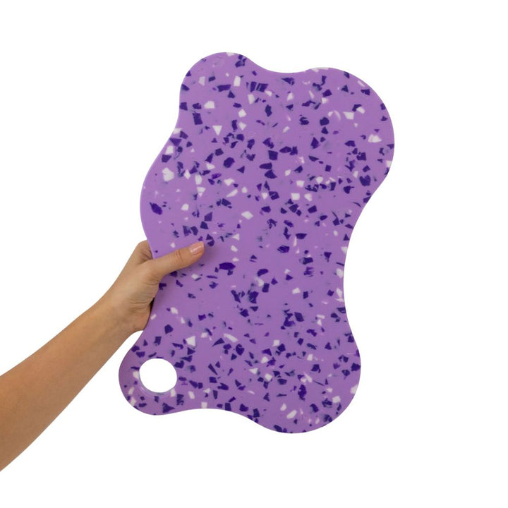 A hand holding up the Nibble and Rest Plum Mama chopping board, showcasing its large purple and redorange speckled design and hanging hole