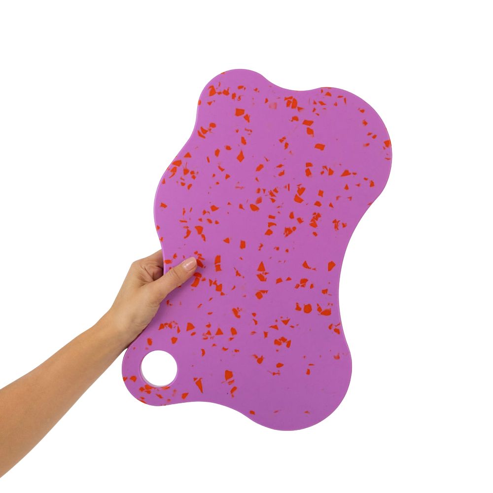 A hand holding up the Nibble & Rest Raspberry Mama chopping board, showcasing its large purple and red orange speckled design and hanging hole