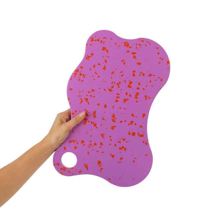 A hand holding up the Nibble & Rest Raspberry Mama chopping board, showcasing its large purple and red orange speckled design and hanging hole